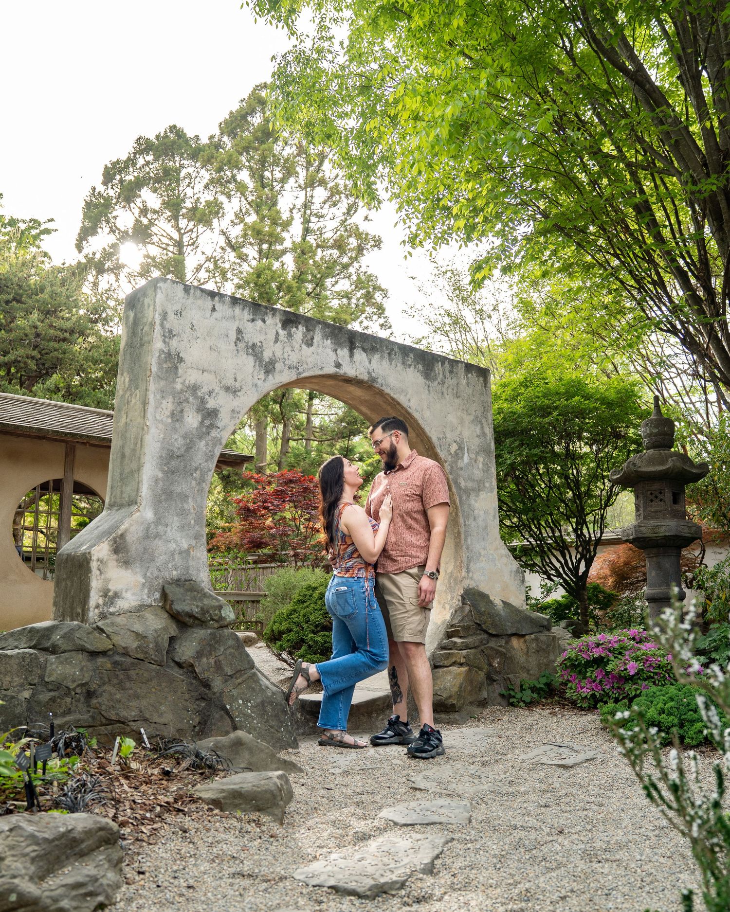 Couple sharing a romantic kiss under a stone moon gate arch in a lush Japanese garden with greenery.