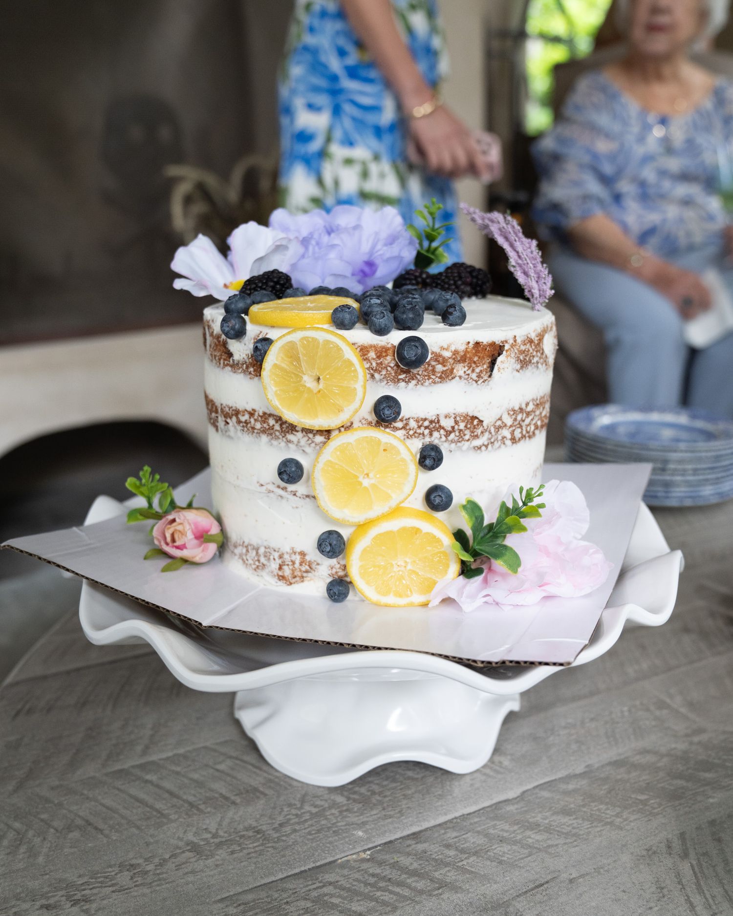 Elegant naked cake decorated with lemon slices, blueberries, and flowers on a white ceramic cake stand.