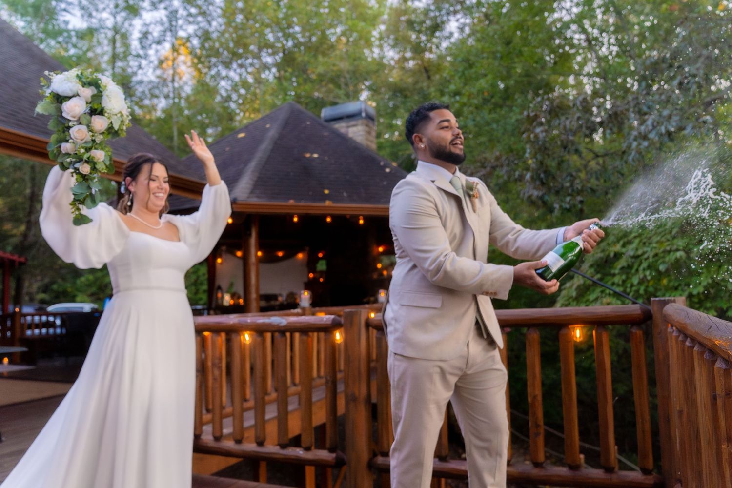 Joyful newlyweds celebrating on wooden deck as groom pops champagne bottle at outdoor wedding venue.