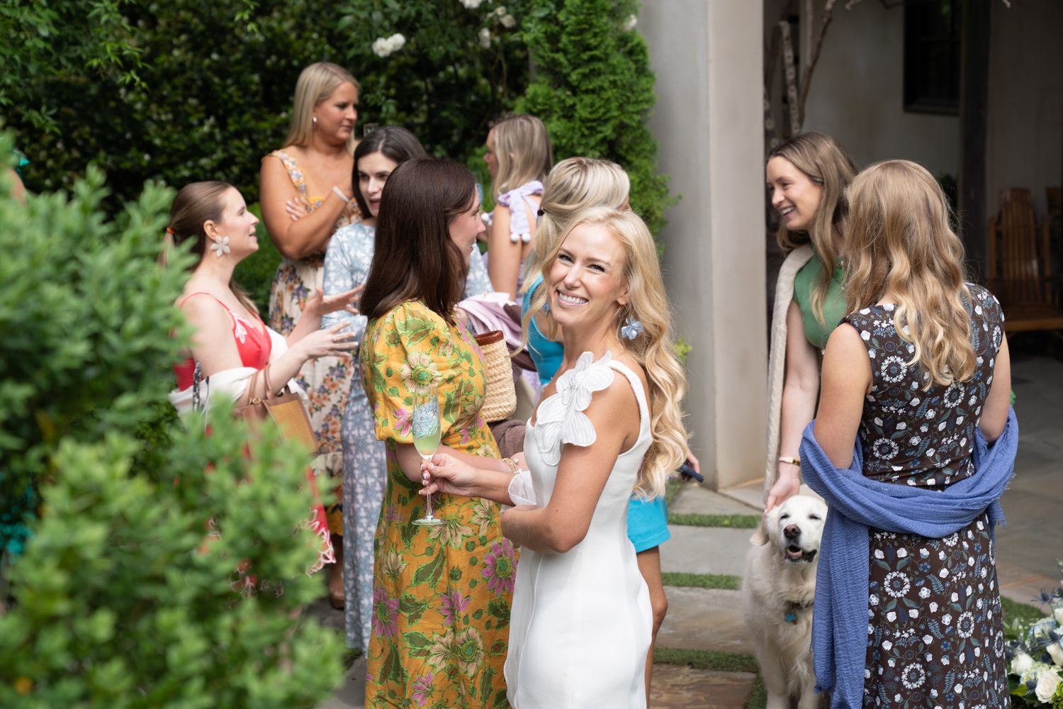 Group of women in elegant dresses socializing outdoors at a garden party with a white dog nearby.