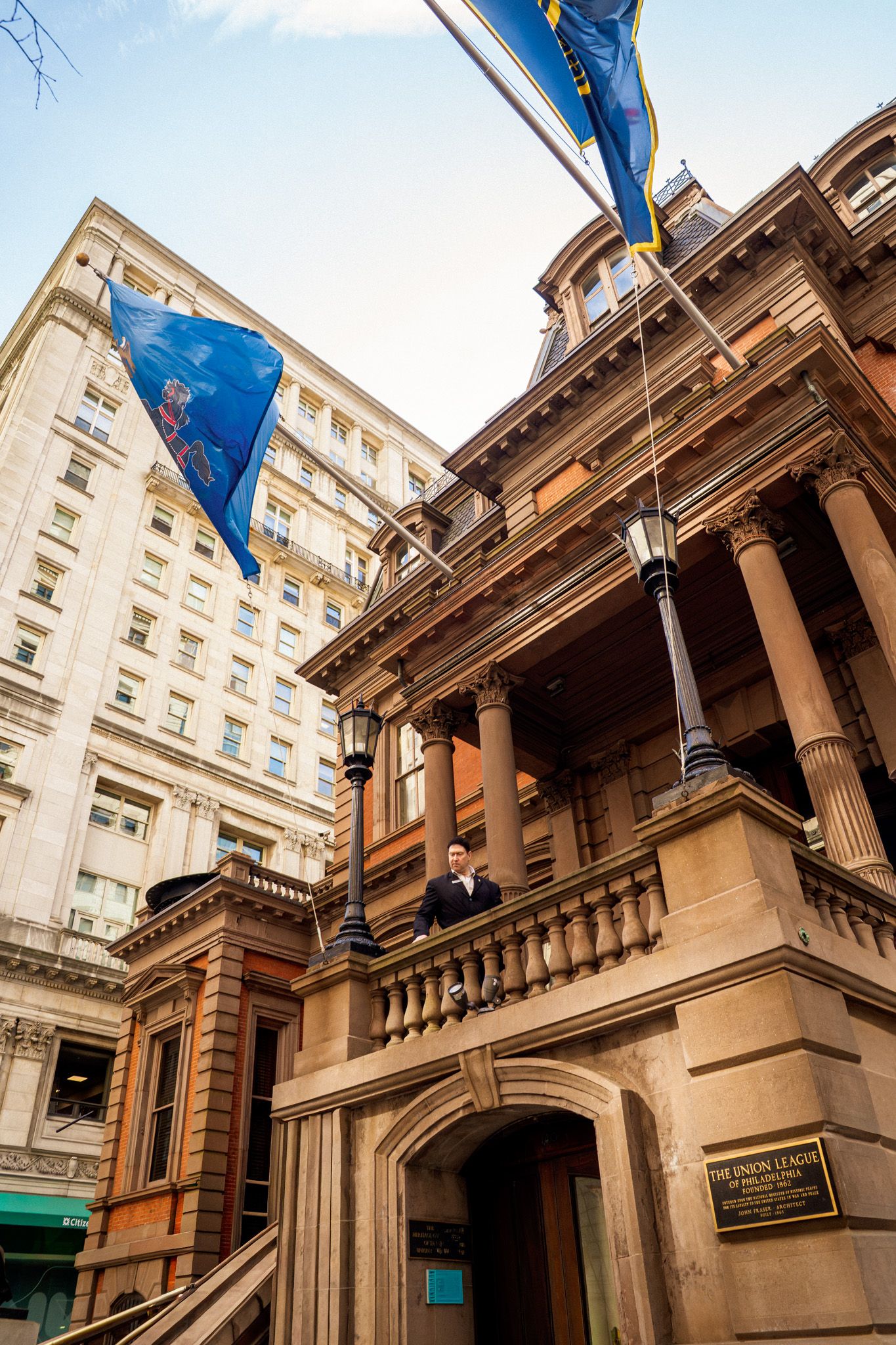 Historic stone building with blue flags, ornate columns, and a balcony in an urban setting.