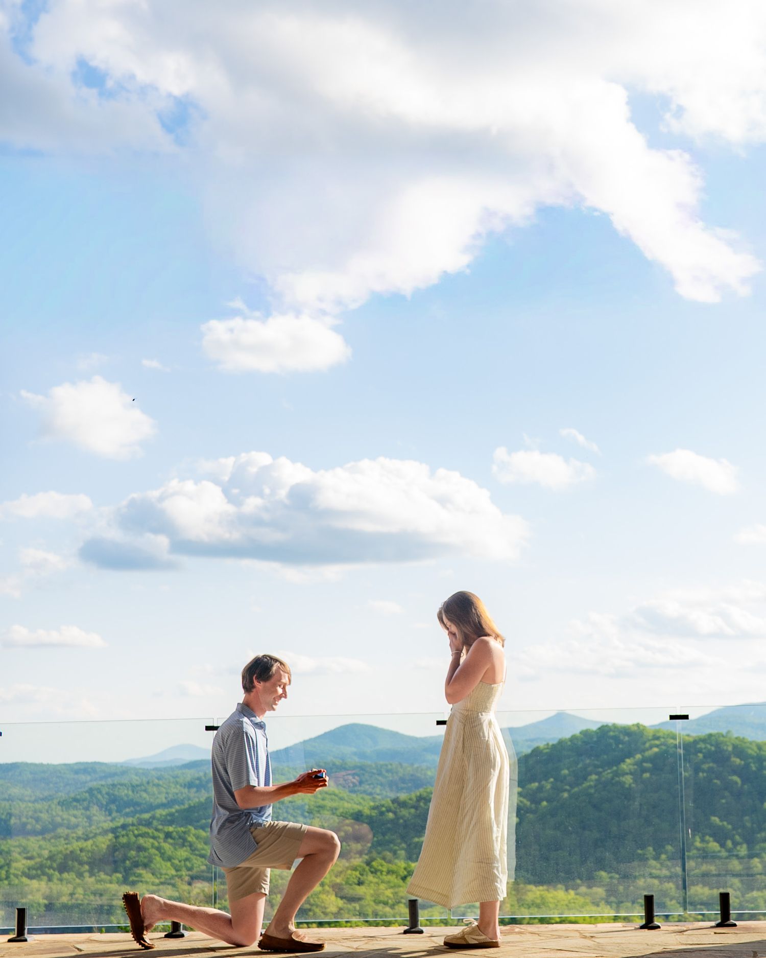 Man proposing on one knee to woman on scenic overlook with rolling green hills and blue sky backdrop.