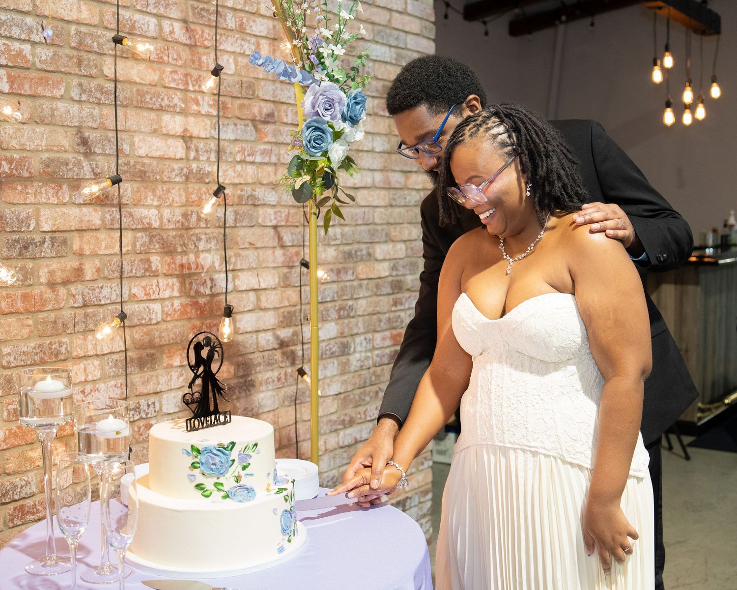 Couple cutting wedding cake at reception, bride in white gown, groom in suit, floral backdrop, brick wall venue.