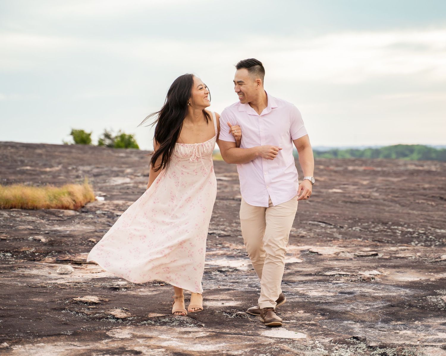 A couple in soft pink and beige outfits shares romantic moments on a rocky landscape during sunset.
