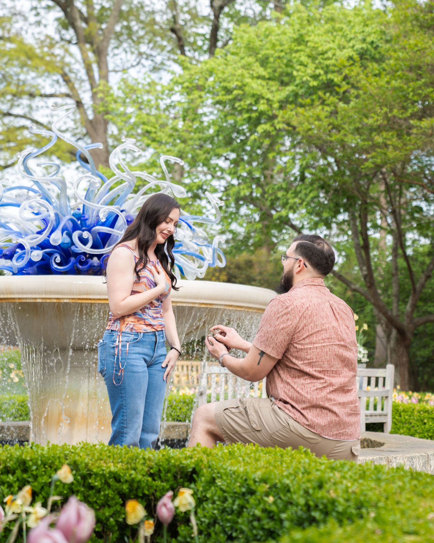 Man proposes to woman on one knee in a garden near a blue glass sculpture fountain surrounded by lush greenery.