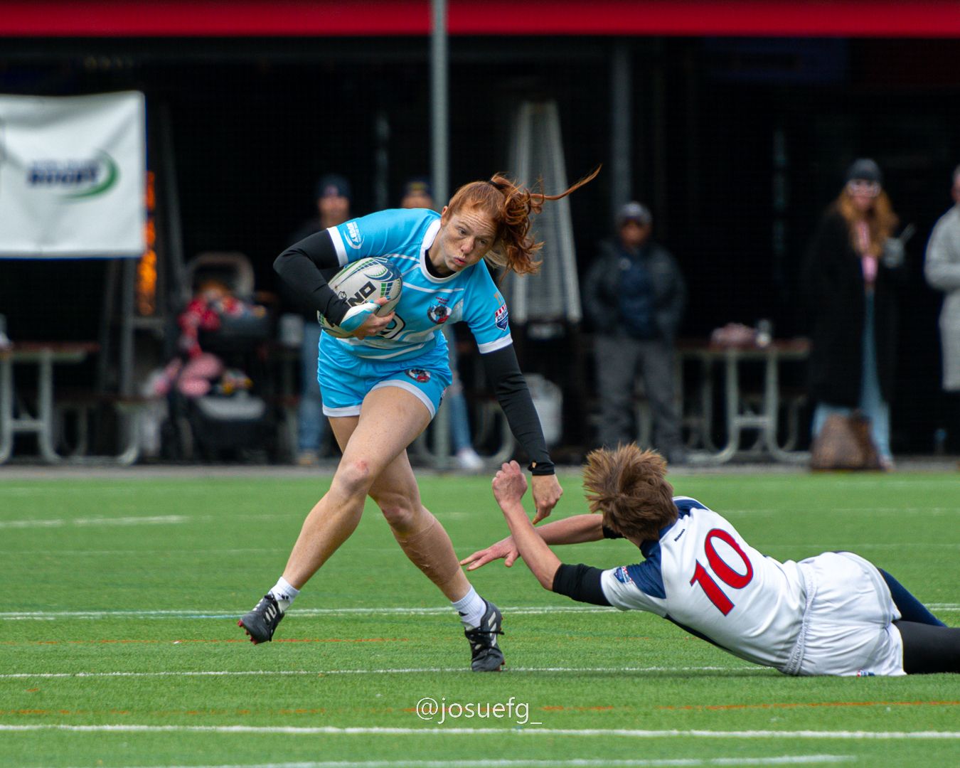 Female rugby player in teal uniform evades diving defender wearing number 10 jersey during competitive match.