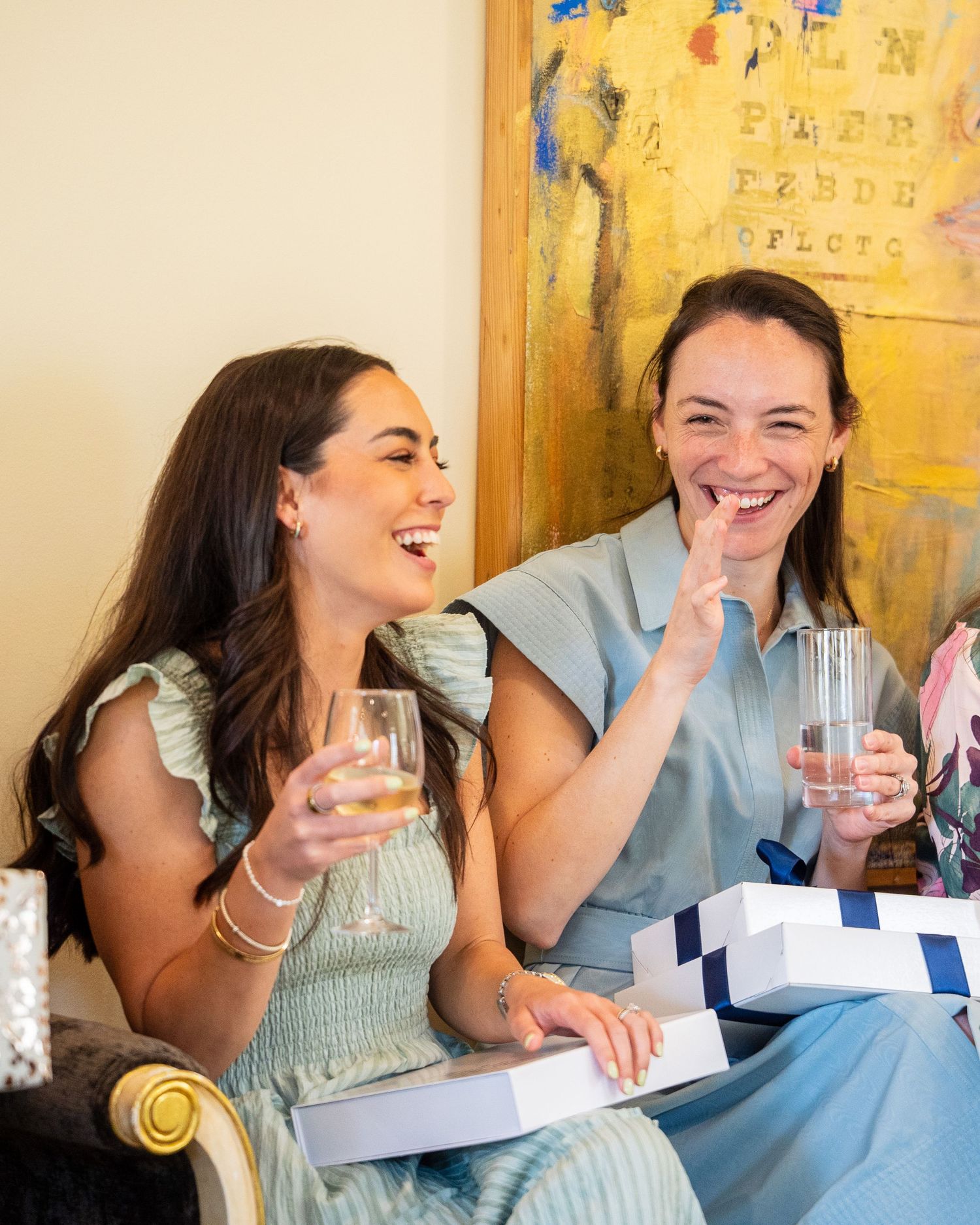 Two women laughing joyfully while holding drinks and opening gift boxes at an indoor celebration.