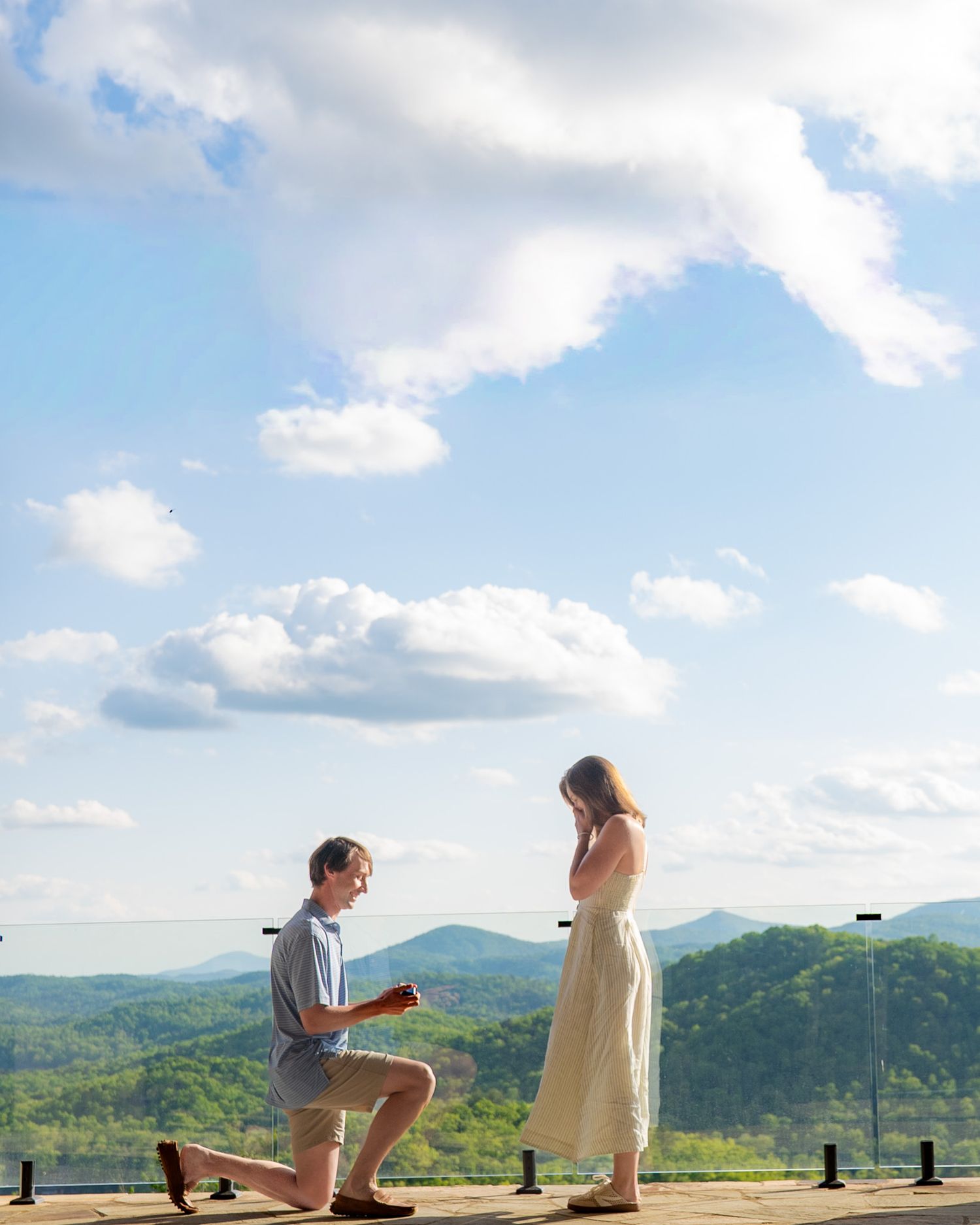 Man proposing on one knee to woman on scenic overlook with rolling green hills and blue sky backdrop.