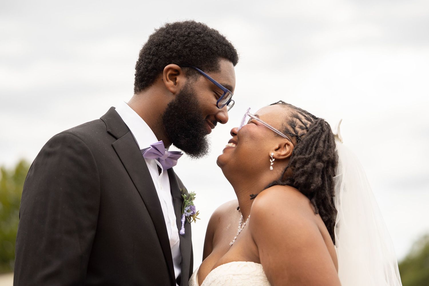 A joyful Black bride and groom share an intimate moment, nearly kissing, dressed in wedding attire outdoors.