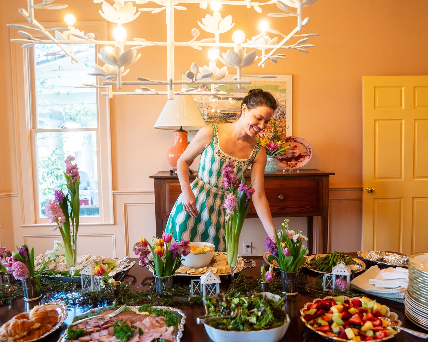 Woman in green checkered dress arranges colorful food spread on decorated table under floral chandelier.