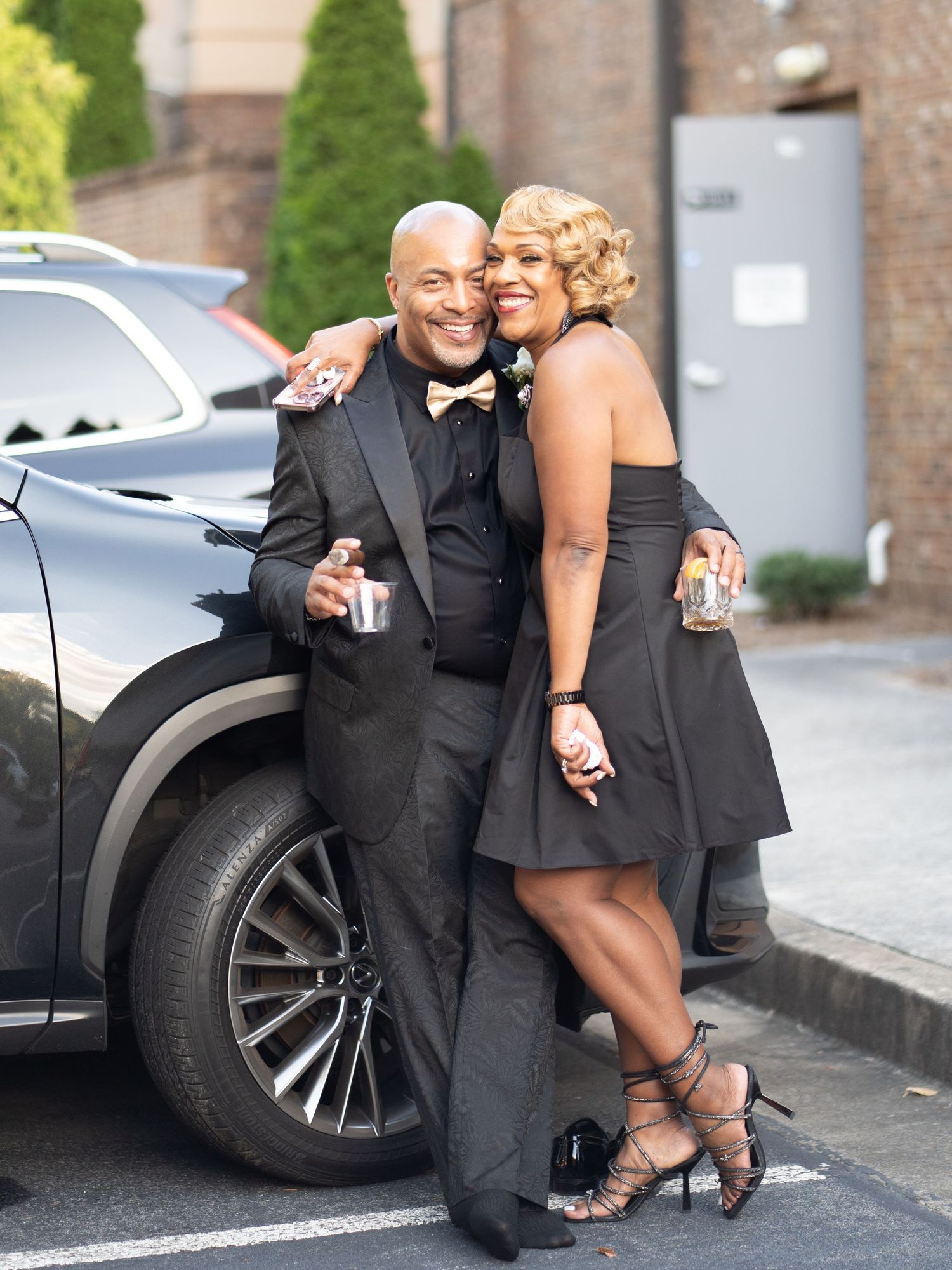 Couple in formal attire poses playfully by luxury car in parking lot.