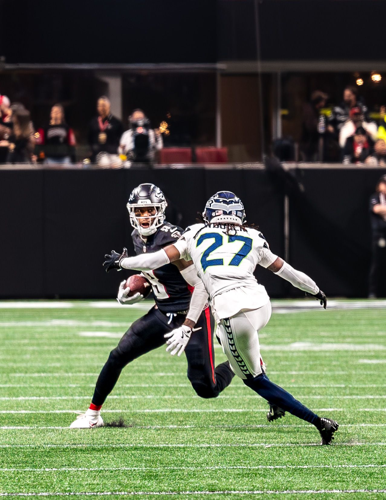 NFL player #27 in Seahawks uniform defends against a receiver during a night game on a green turf field.