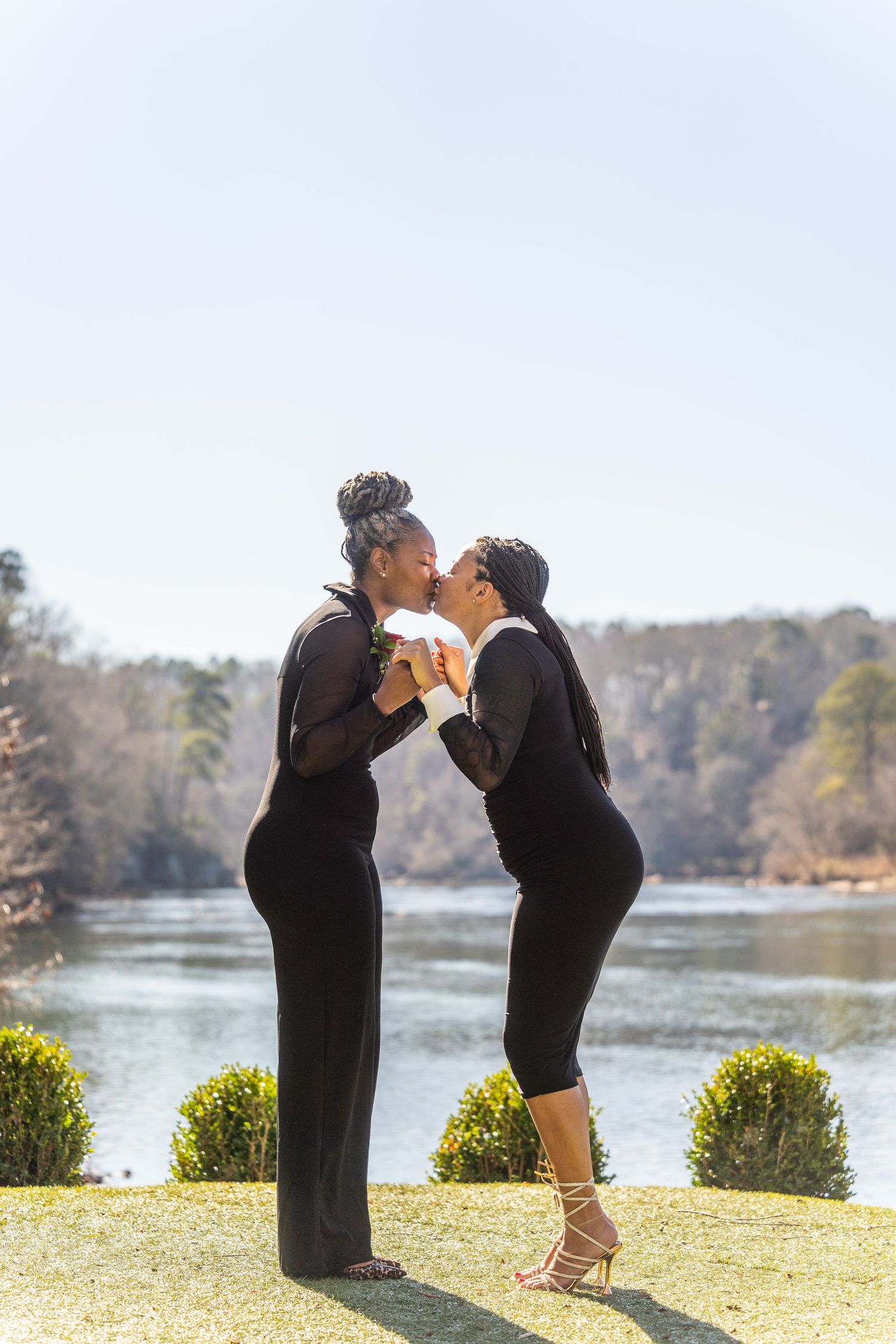 Two women in elegant black outfits share a kiss by a serene lakeside on a sunny day.