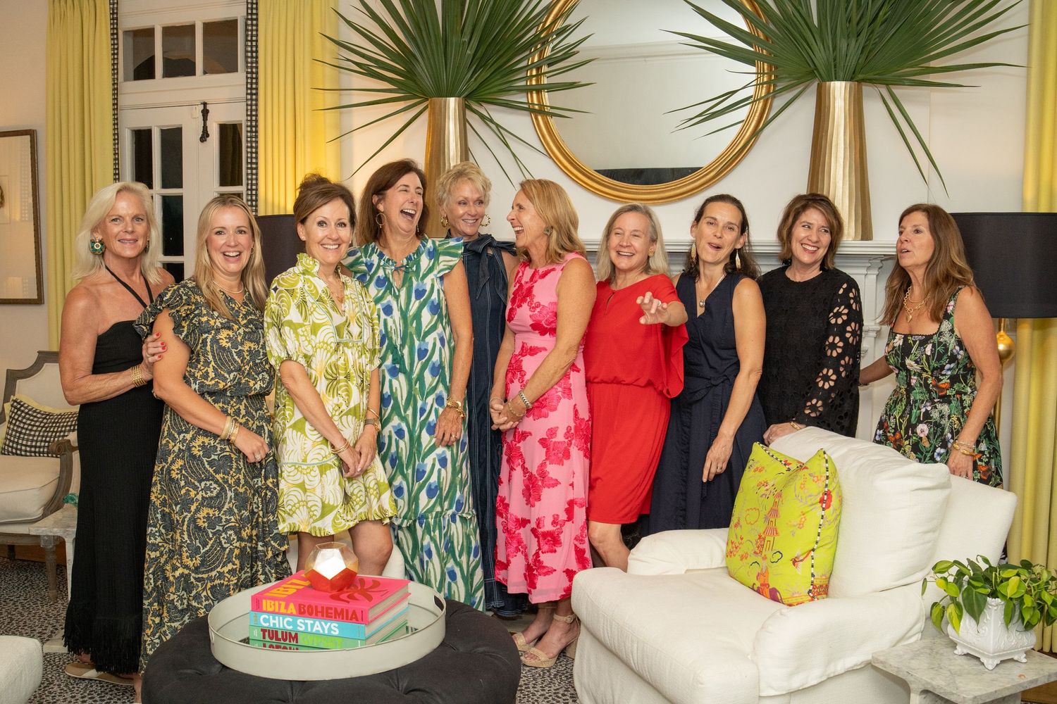 Group of ten elegantly dressed women smiling together at an indoor party near a white sofa.