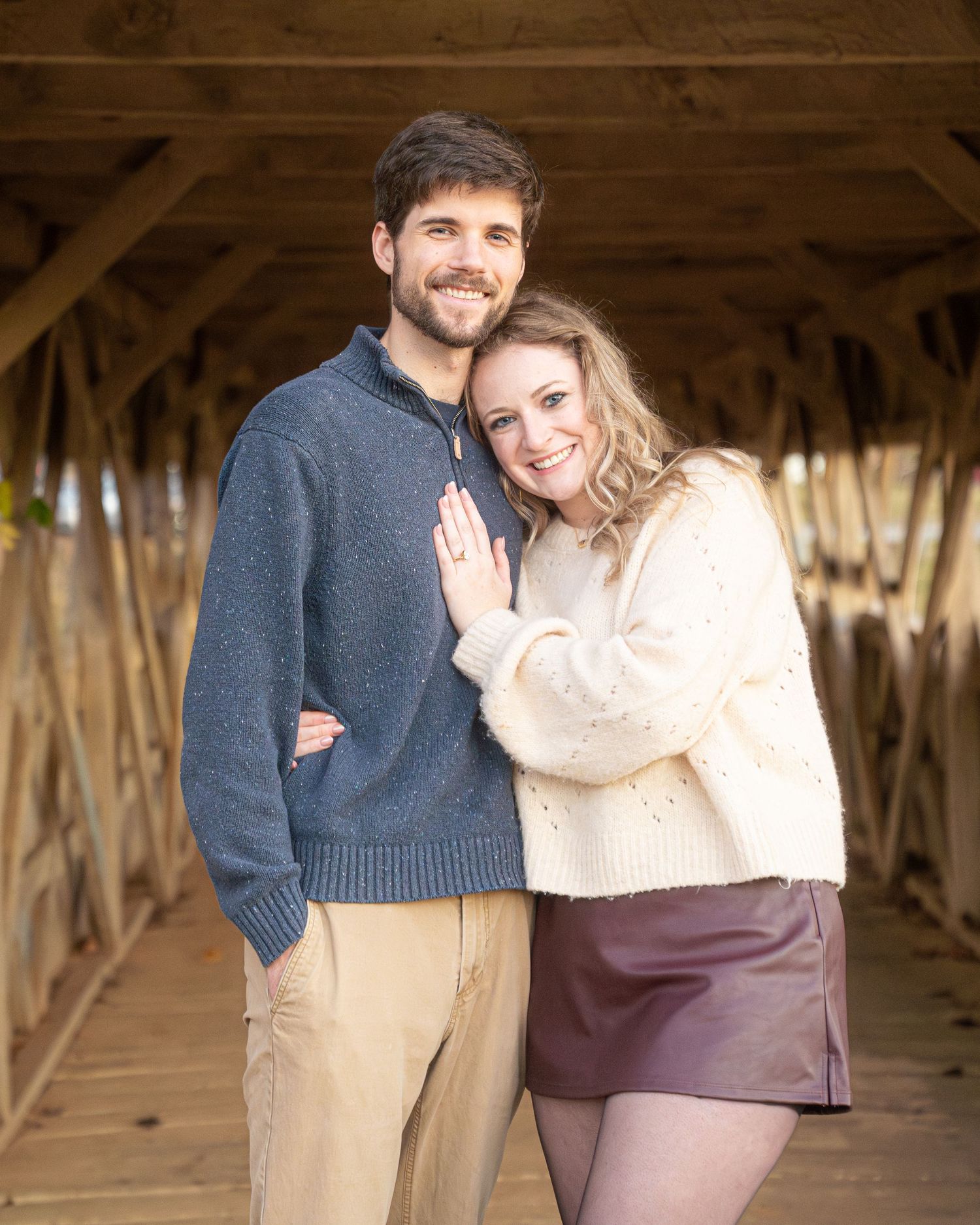 A couple poses together inside a covered wooden bridge wearing cozy sweaters.