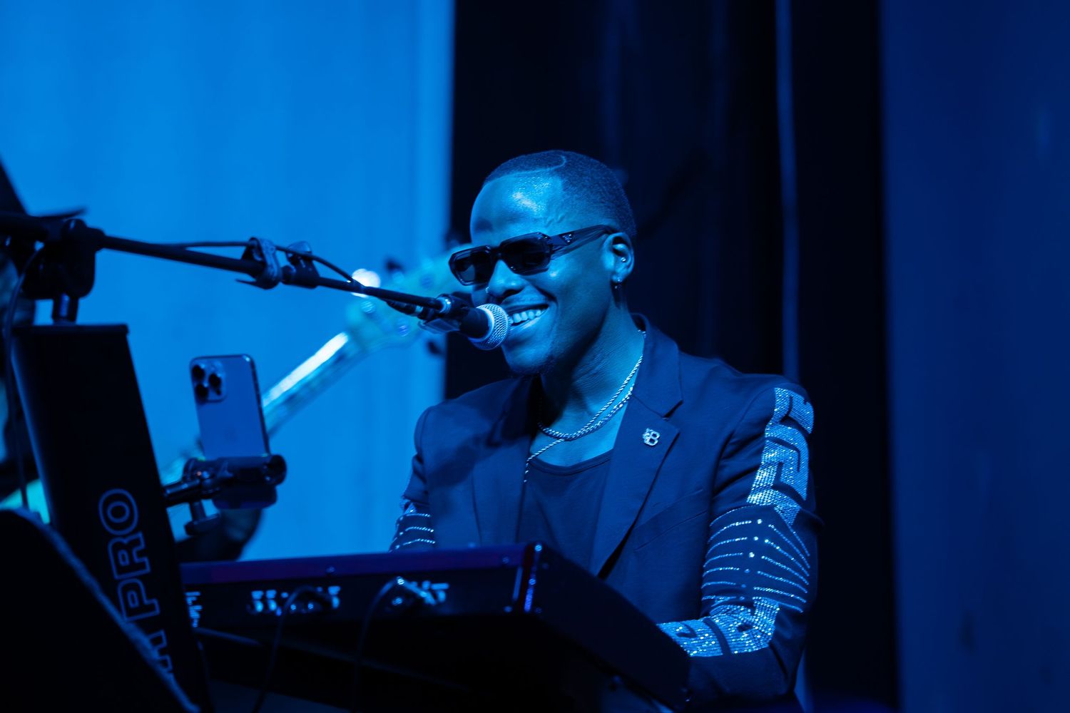 A smiling musician wearing sunglasses performs at a keyboard under blue stage lighting.