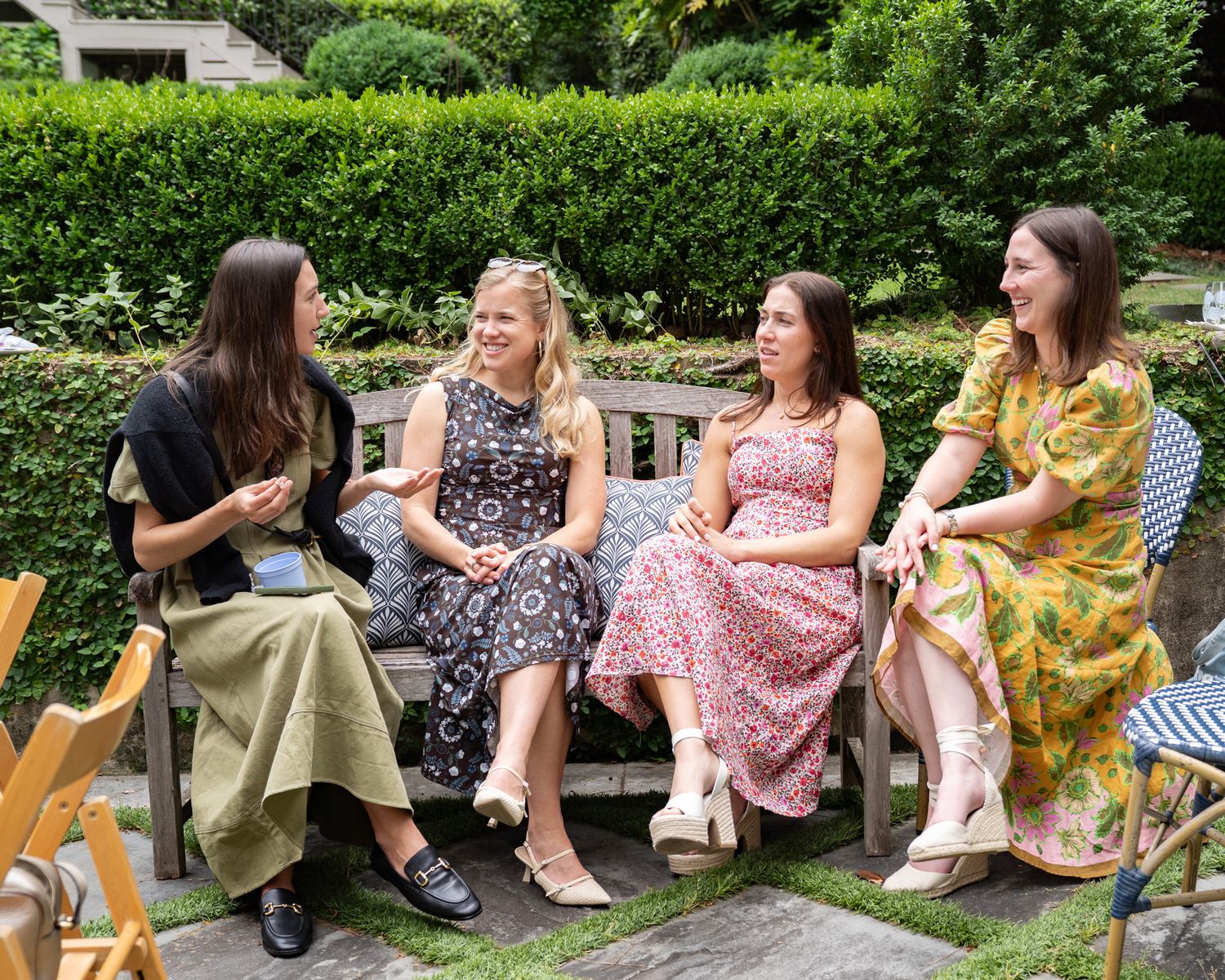 Four women in floral summer dresses sit on a garden bench, laughing and conversing outdoors.