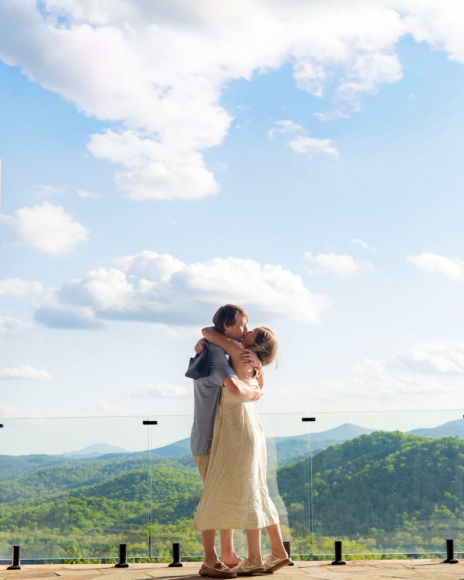 Couple embracing and kissing on a scenic overlook with lush green mountains and blue sky backdrop.
