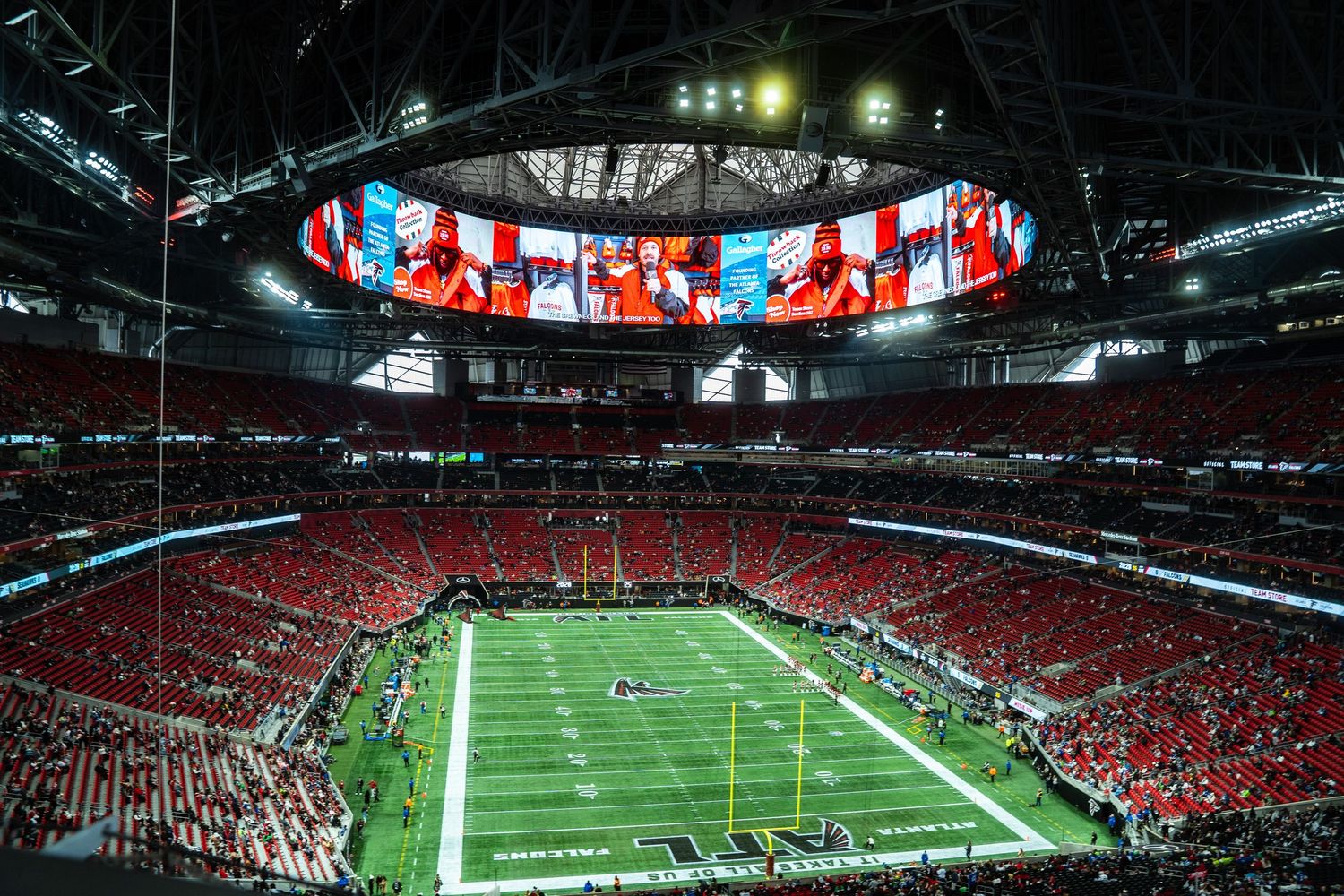 Aerial view of a packed NFL stadium with a massive halo video board display and green field during a game.