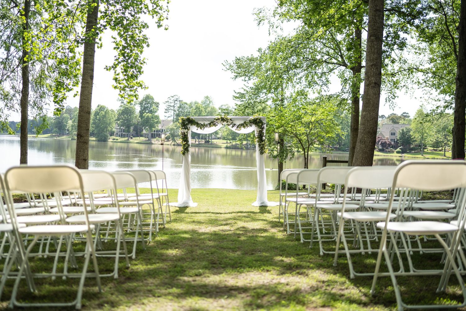Outdoor wedding ceremony setup with white folding chairs, floral arch, and serene lake backdrop surrounded by lush green trees.