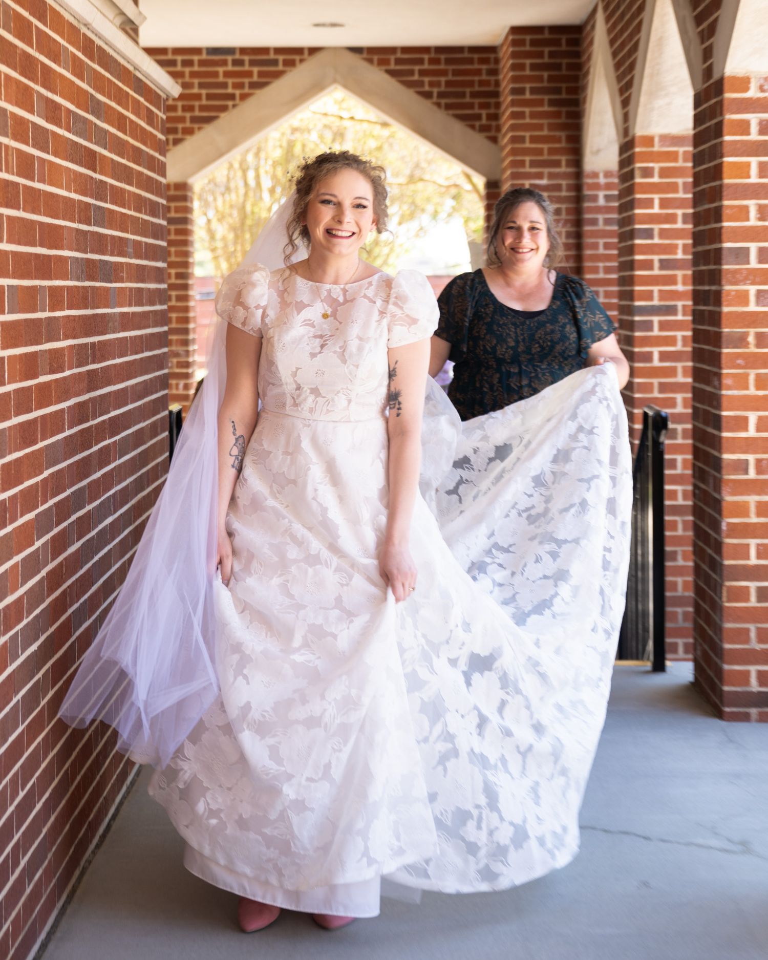 Bride in lace wedding gown and veil smiles as woman in dark dress holds her train outside brick church.
