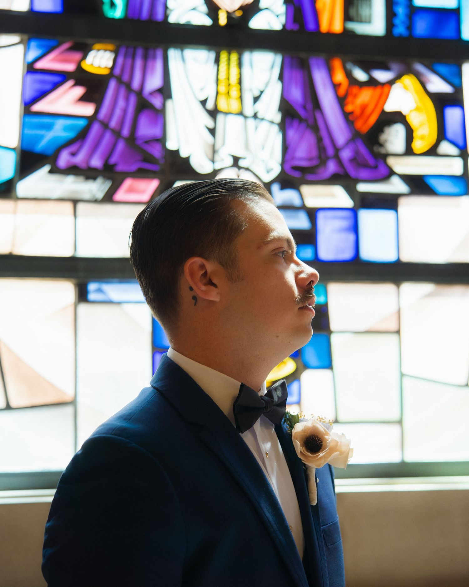 Groom in blue suit and bow tie with boutonniere stands before colorful stained glass window in church.