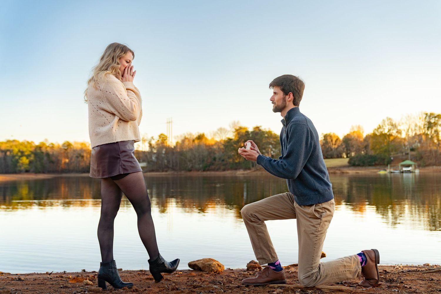 Man proposes to woman on one knee by a lake at sunset during autumn, holding a ring box.
