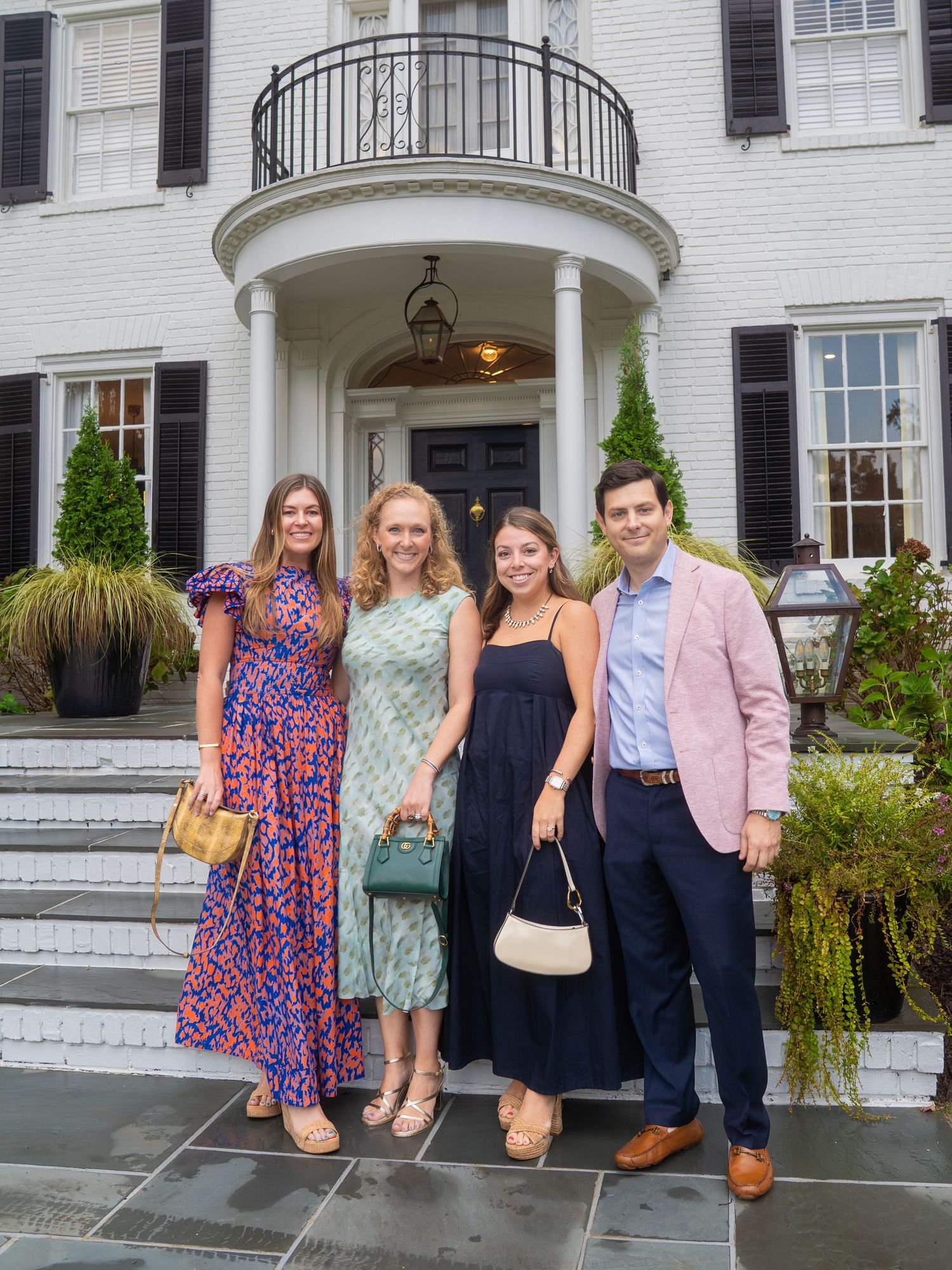Four elegantly dressed friends pose together on steps outside a grand white colonial mansion with black shutters.