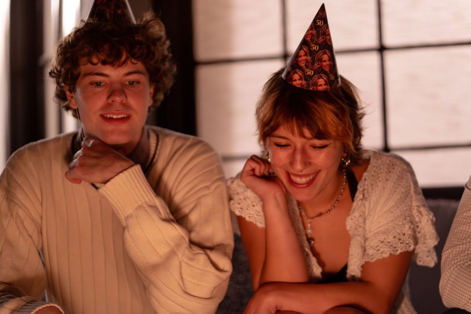 Two young people wearing festive party hats smile and laugh together at a celebration party.