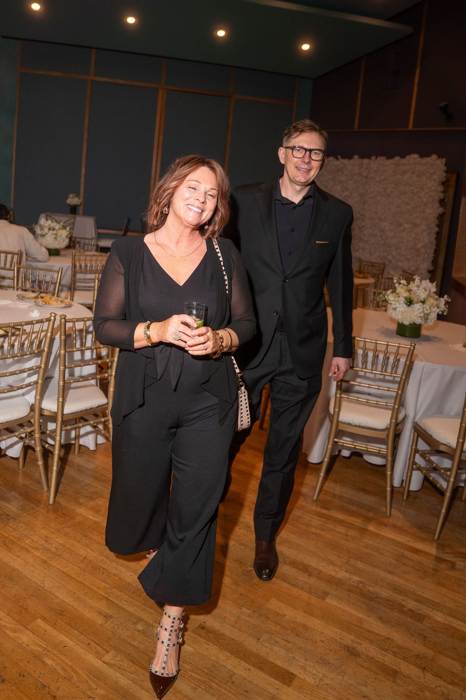 Couple dressed in black attire smiling and walking through an elegant event venue with gold chairs.