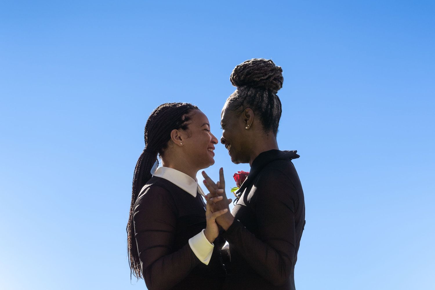Two women in black outfits share a romantic moment under a clear blue sky, one holding a red rose.