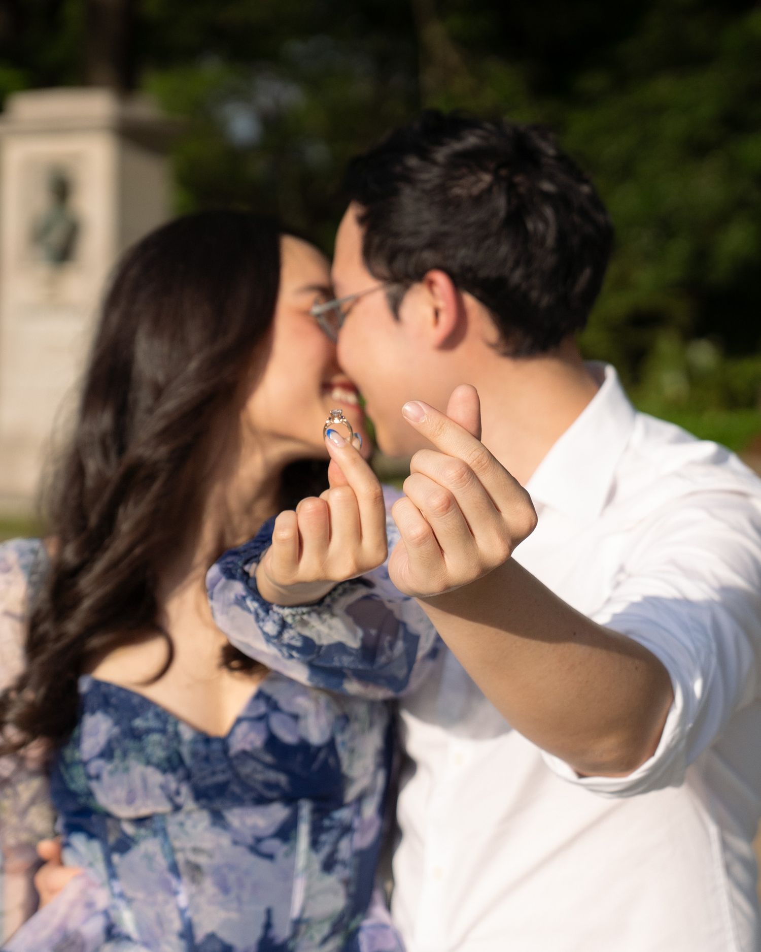 A couple shares a kiss while showing off an engagement ring in a sunny outdoor park setting.