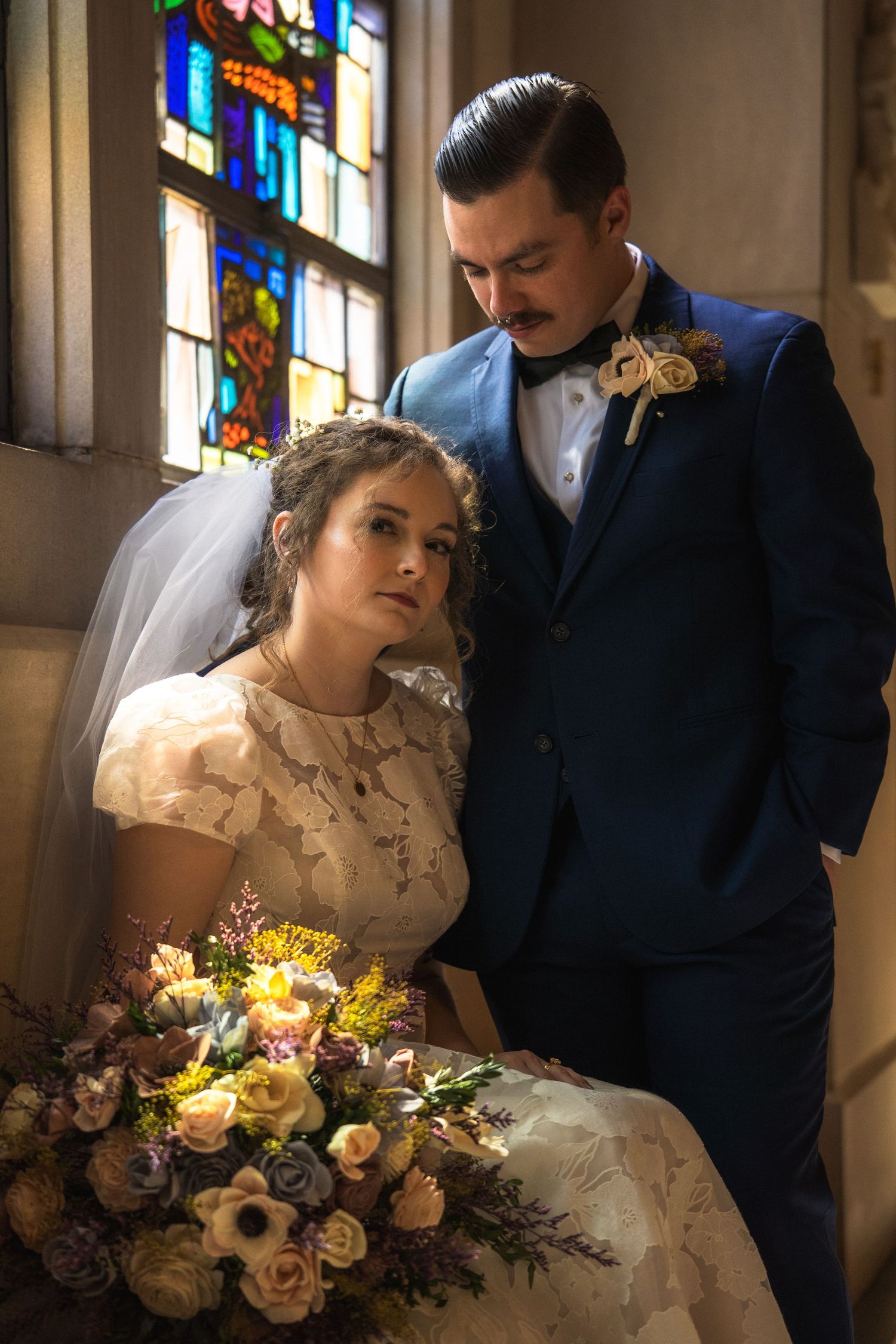 Bride in lace dress holding floral bouquet sits beside groom in blue suit near stained glass church window.