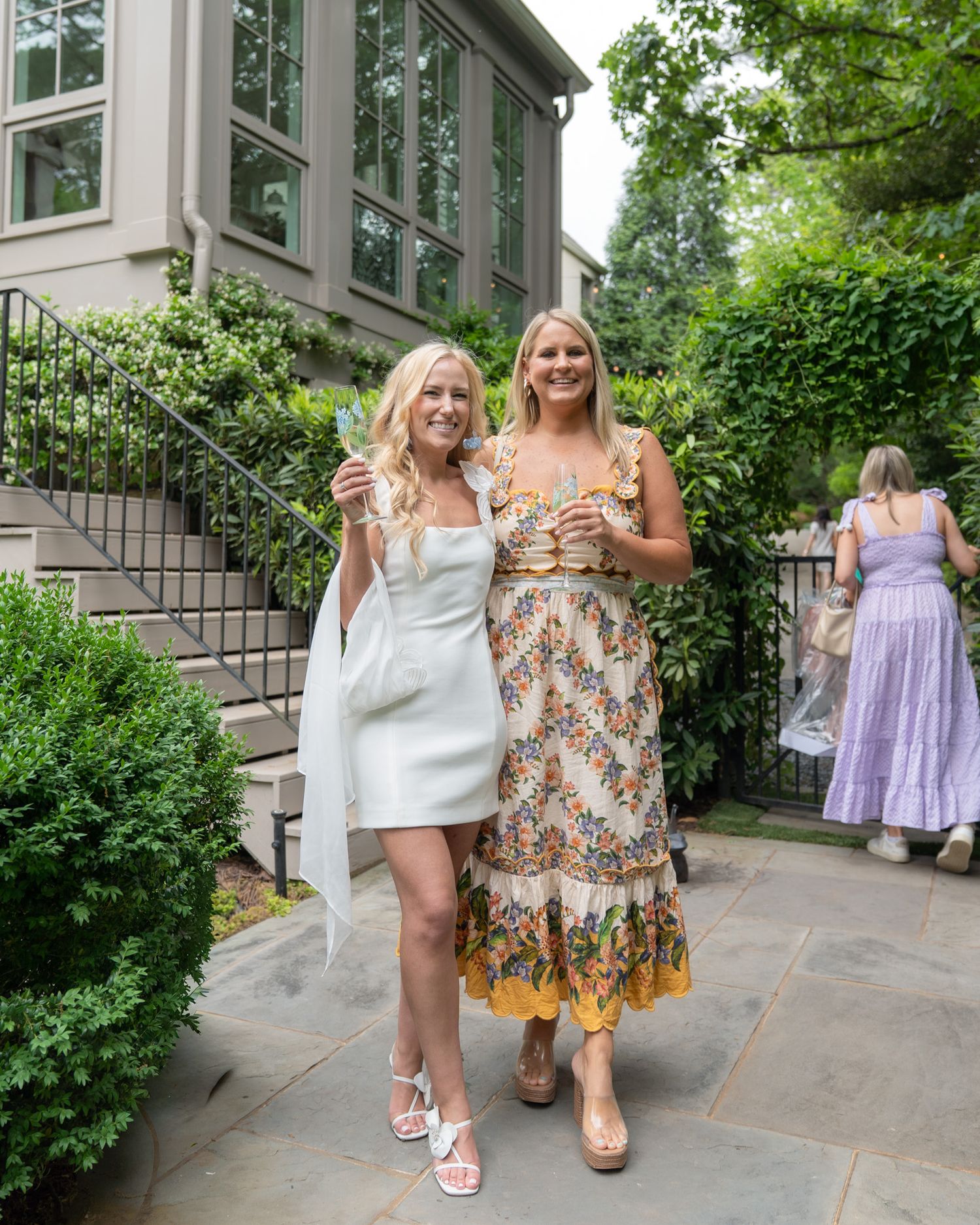 Two smiling women in summer dresses holding drinks at an outdoor garden party event.