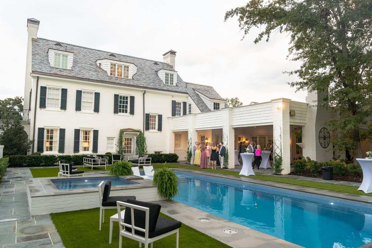 Luxury white estate with pool, black shutters, and elegantly dressed guests gathered for an outdoor evening event.