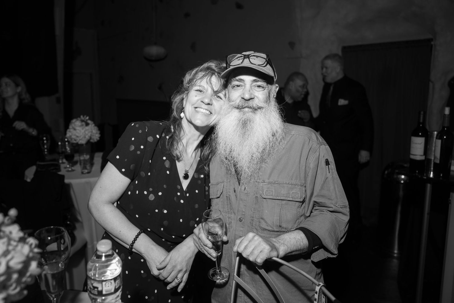 Smiling couple posing together at indoor event, man with long white beard in casual shirt, black and white photo.
