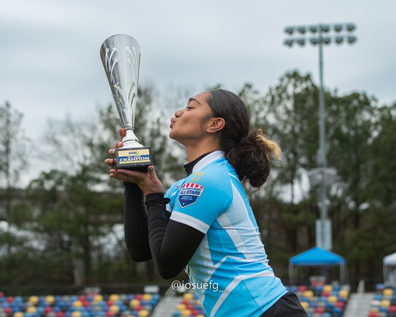 Female athlete in blue jersey kisses trophy on sports field after championship victory.