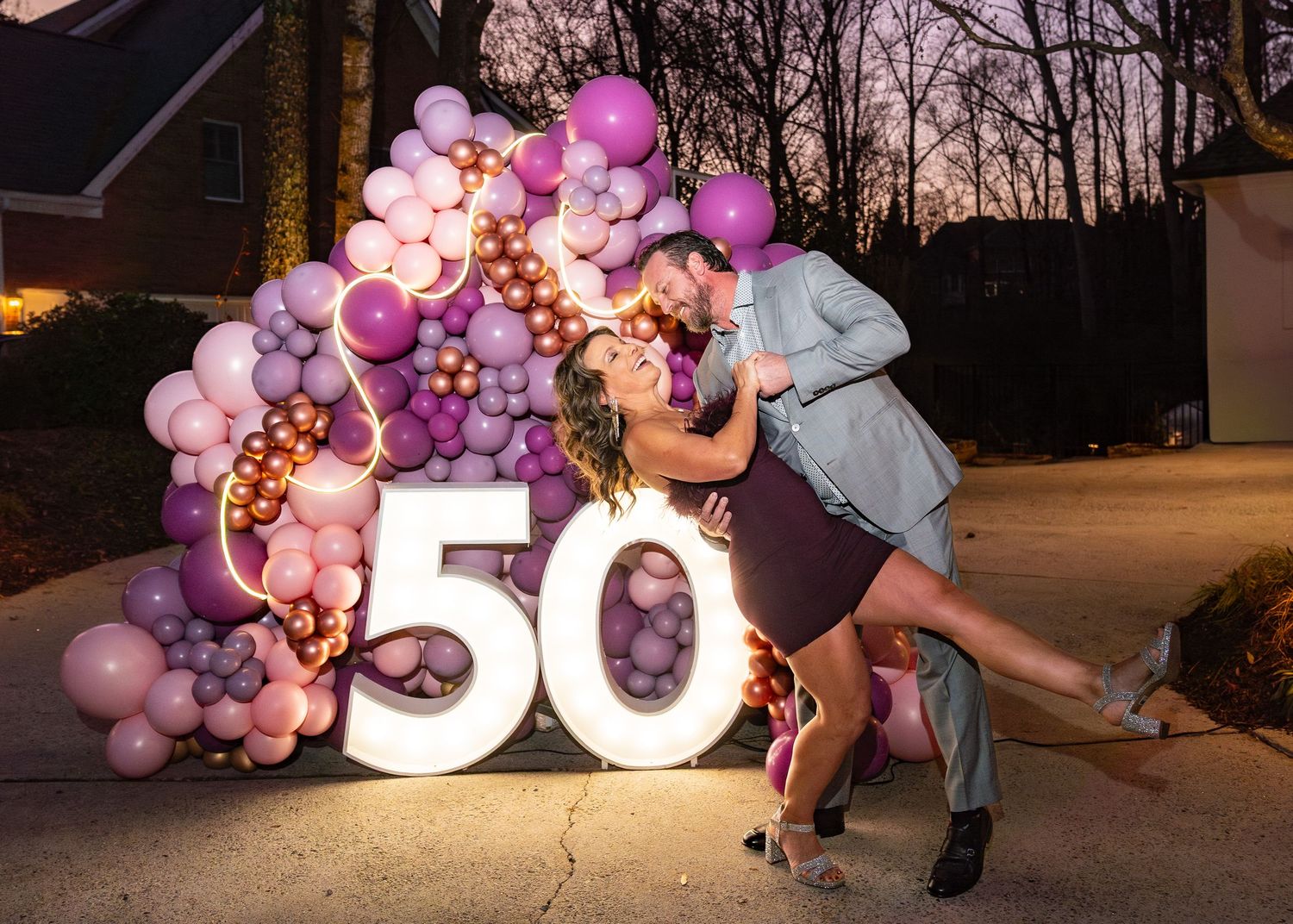 Couple posing romantically beside illuminated 50 balloon display with purple balloons at outdoor birthday celebration.