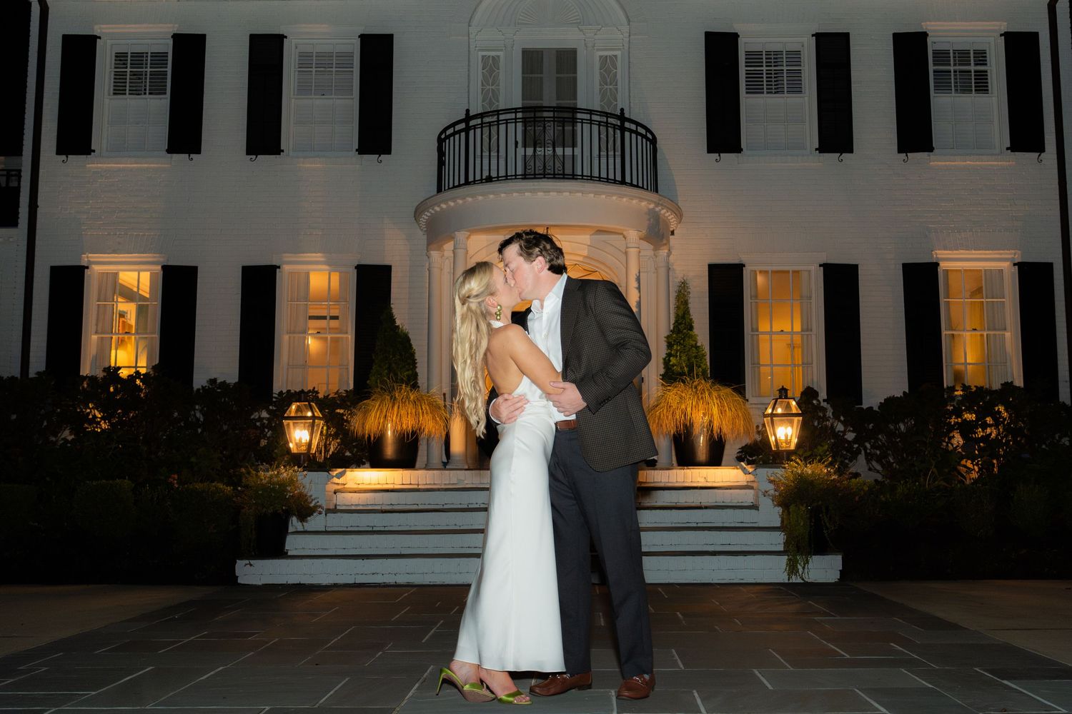 Elegant evening portrait session outside white colonial mansion with lit windows.