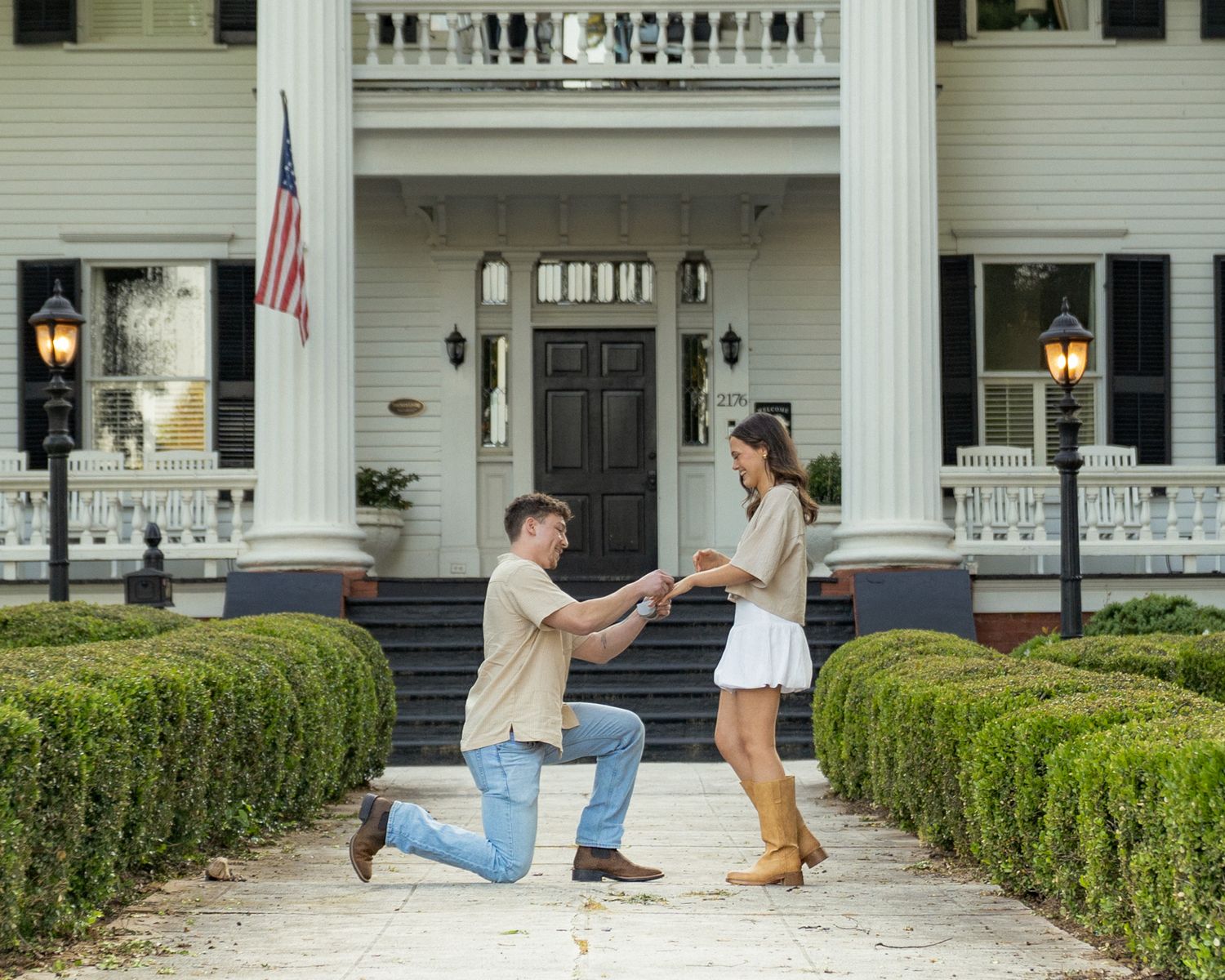 Man proposes on one knee to woman in front of elegant white Southern mansion with columns and American flag.