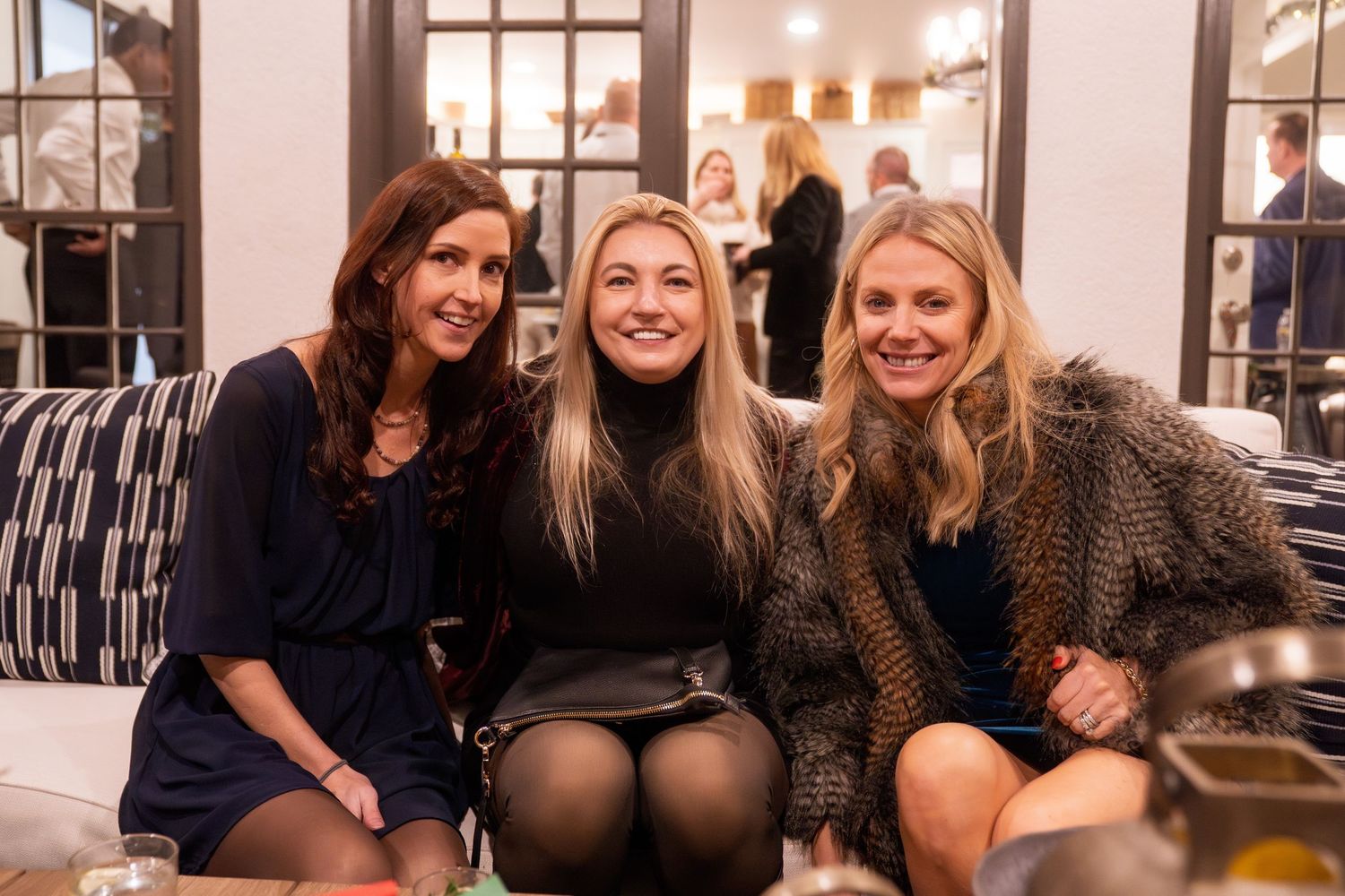 Three smiling women pose together at an indoor social gathering, dressed in stylish outfits in a warmly lit venue.