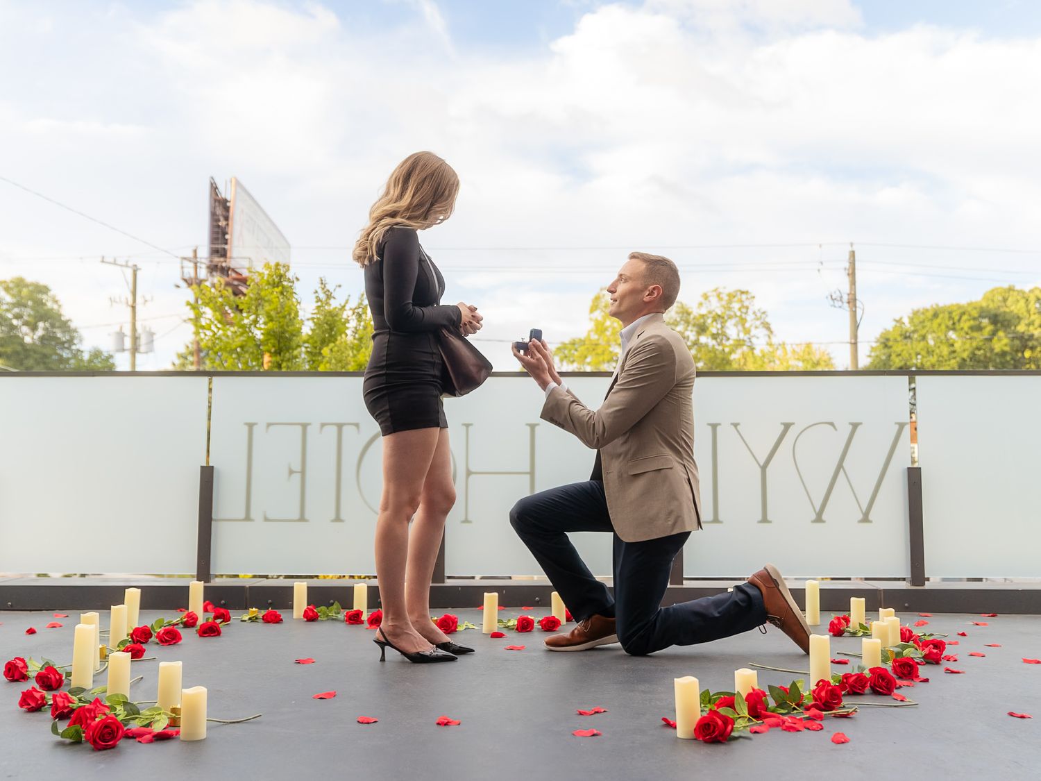 Man proposes to woman on rooftop surrounded by candles, rose petals, and red roses in a romantic heart arrangement.