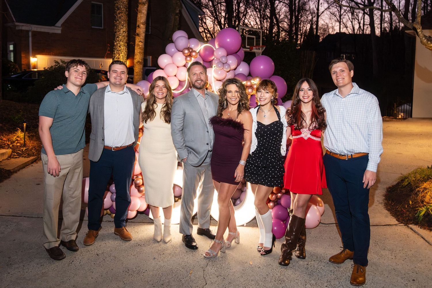 Group of seven people posing together outdoors at dusk near pink balloon decorations at a celebration event.