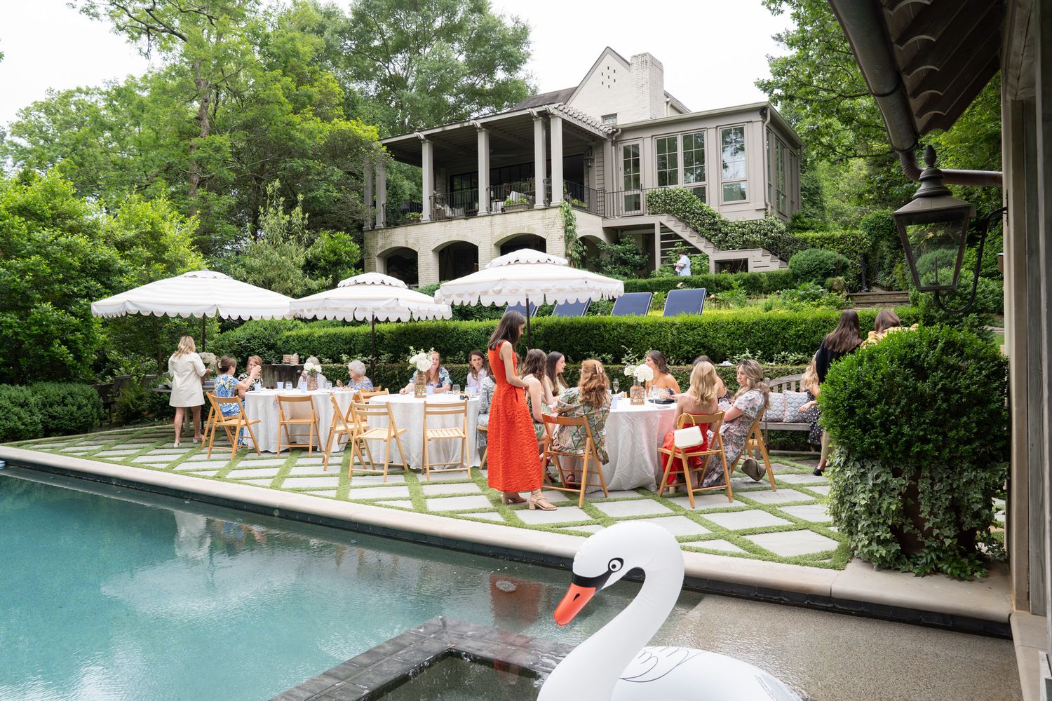 Elegant outdoor poolside garden party with guests dining under white umbrellas beside a luxury home with swan float.