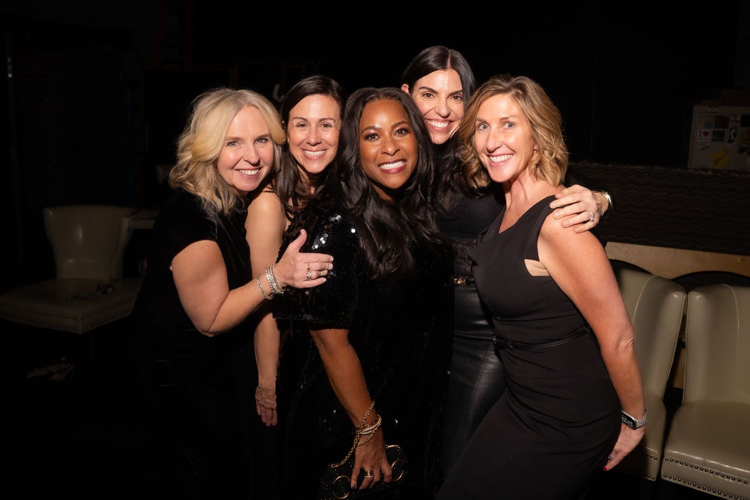 Five women smiling and posing together at an evening social event.