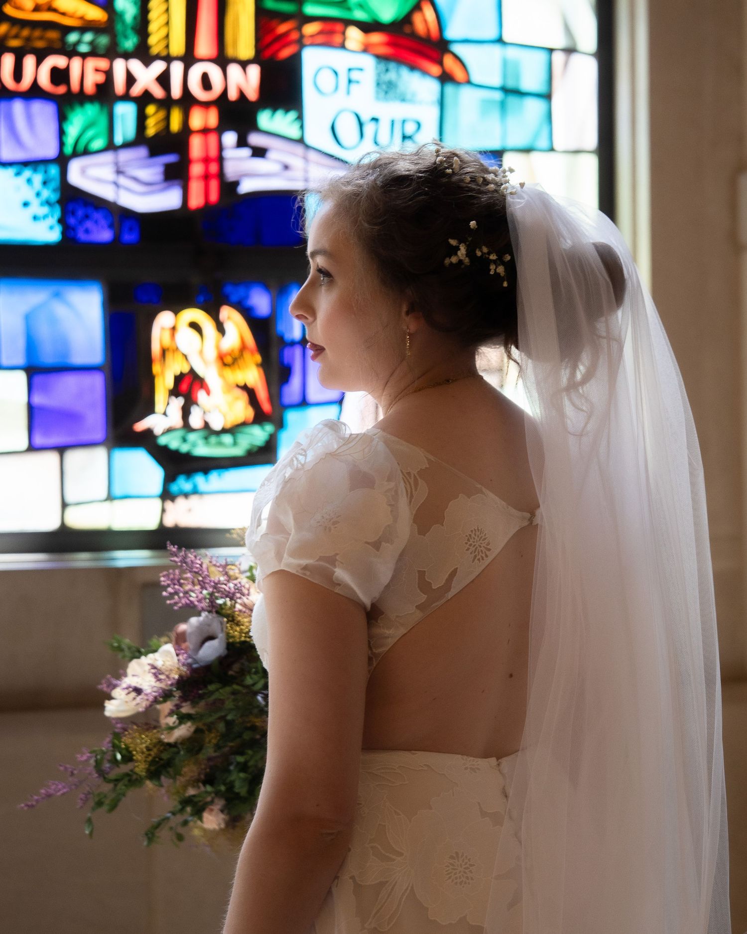 Bride in white gown and veil holds bouquet before colorful stained glass windows in a church.