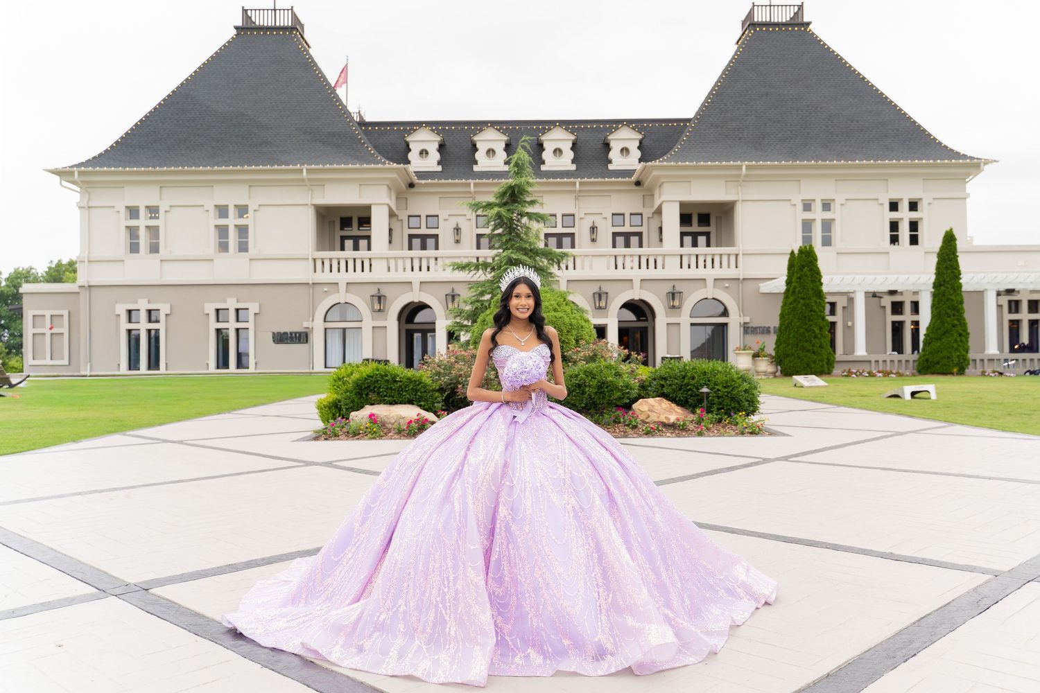 Young woman in lavish lavender ball gown wearing tiara poses in front of elegant white mansion for quinceañera.