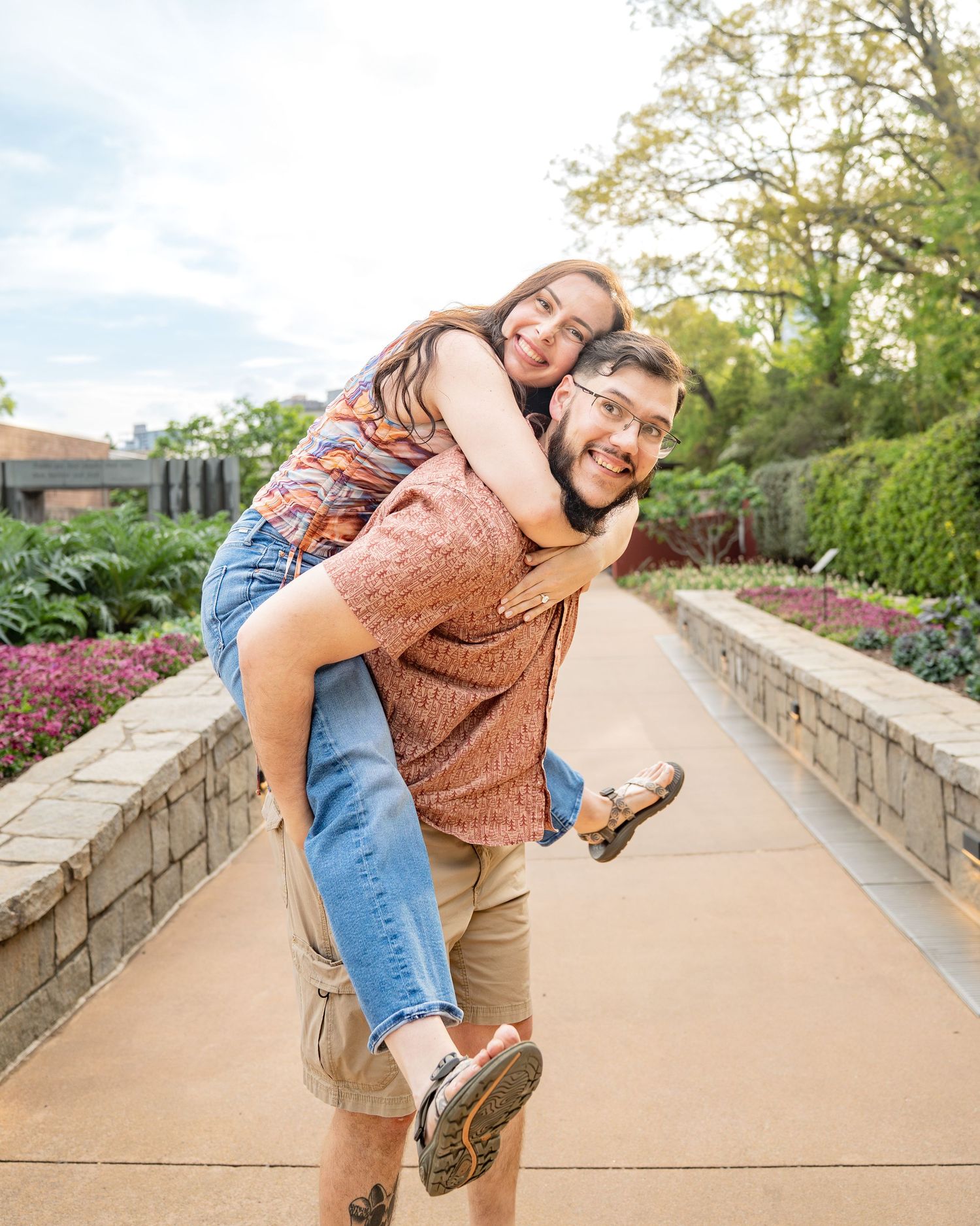 A young couple laughing as the man gives the woman a piggyback ride on a scenic garden pathway.