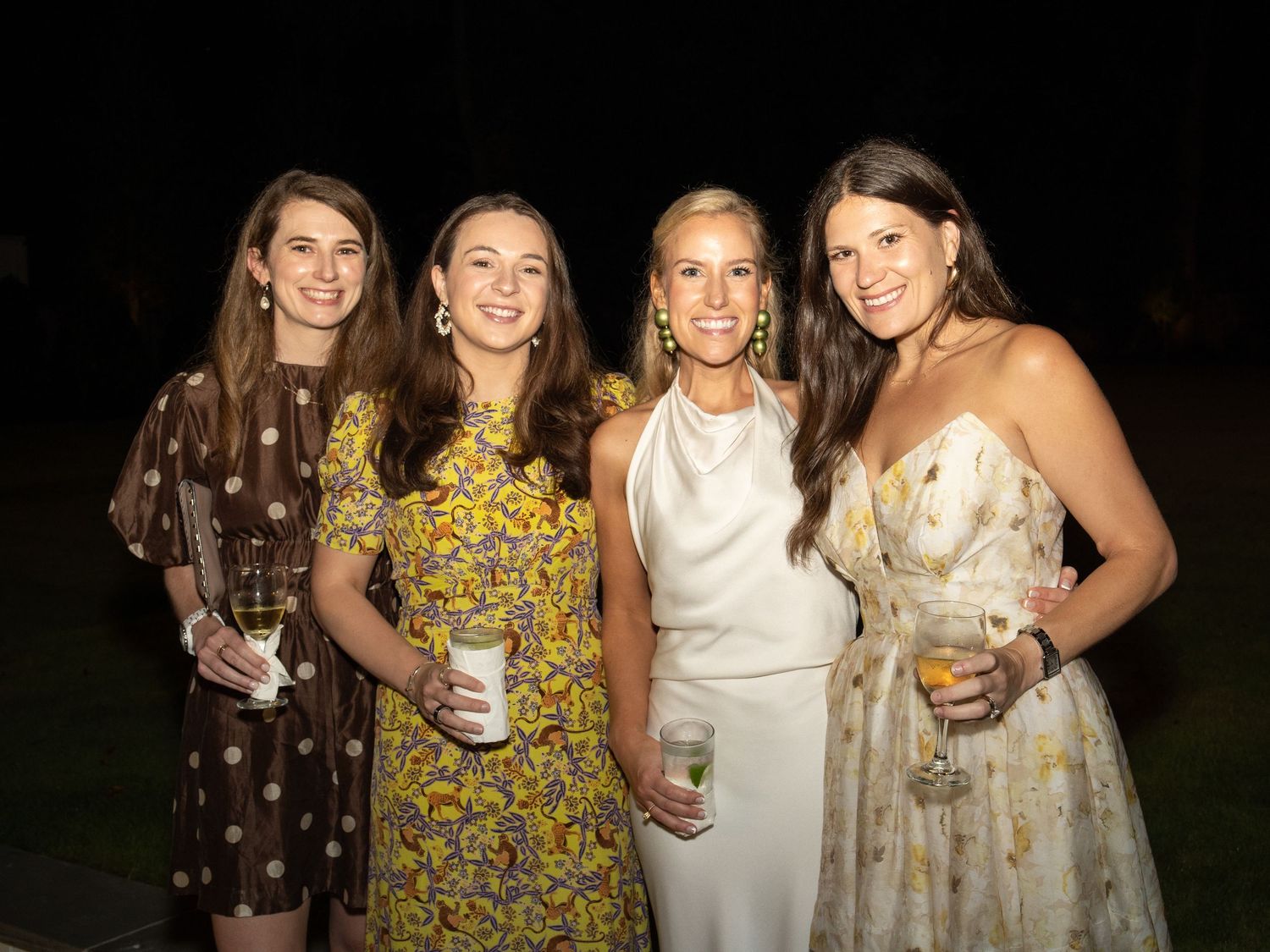 Four young women in floral and white dresses smiling together at an outdoor evening social event.