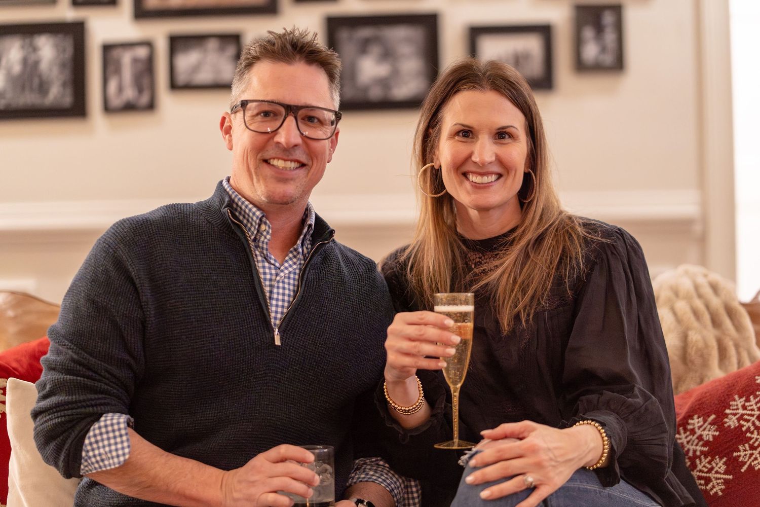 A smiling couple toasting with champagne glasses while sitting together on a couch at a festive holiday gathering.
